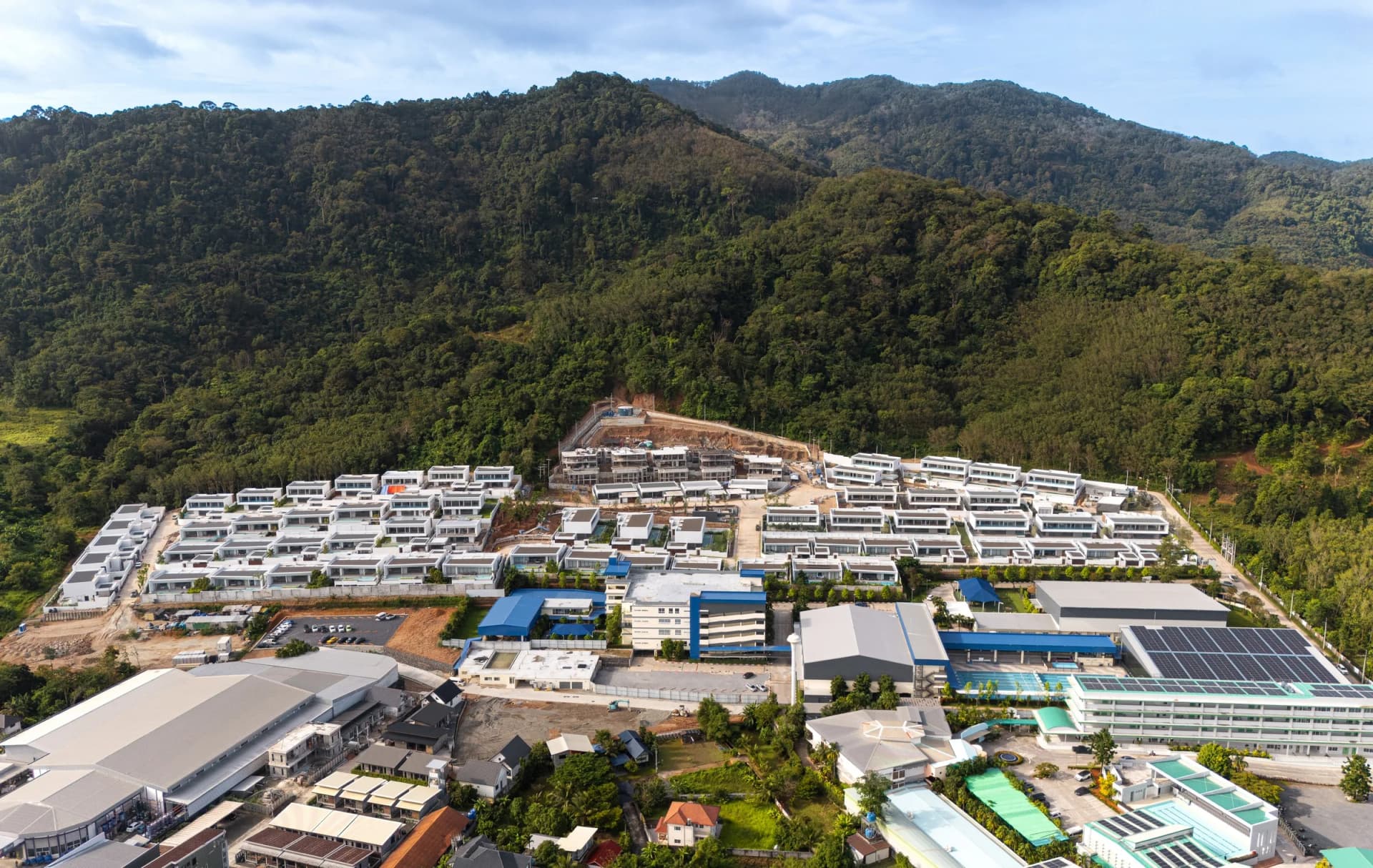 Drone photo from the valley: rows of flat-roof pool villas against the forest, the school buildings and sports hall in front